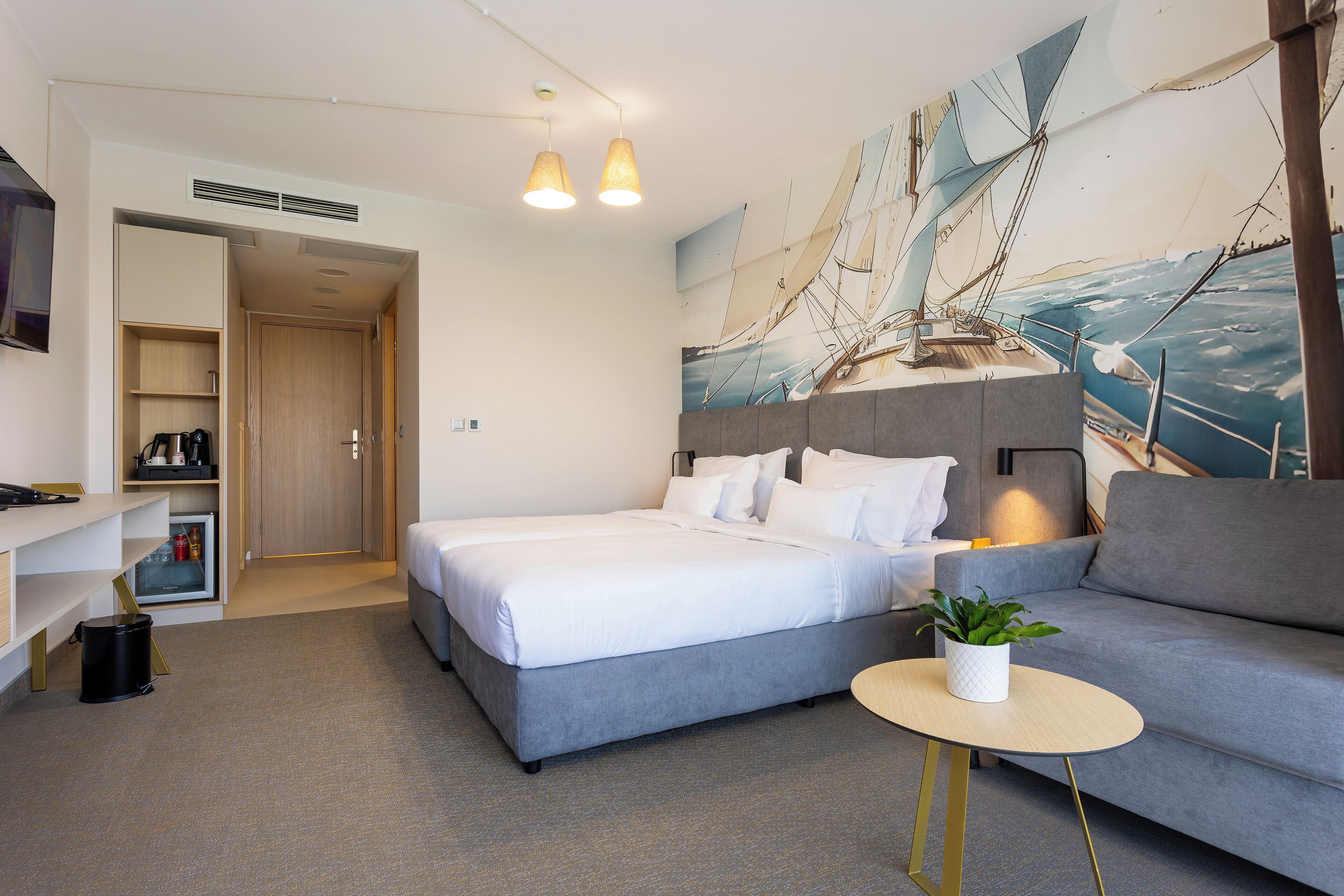 A modern hotel room with a large bed, a sofa, a small table with a plant, and a wall mural of a sailing ship.