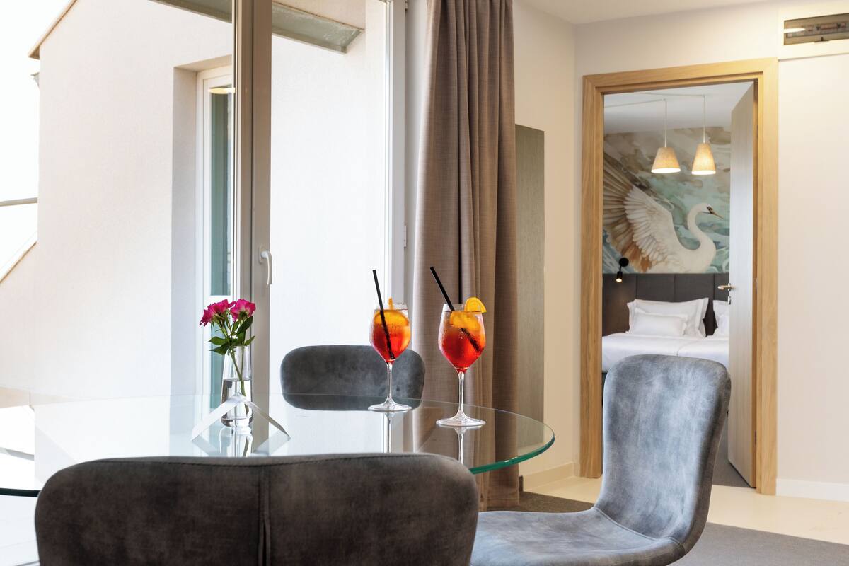 A hotel room with a glass table, two drinks, a chair, and a bed with a painting.