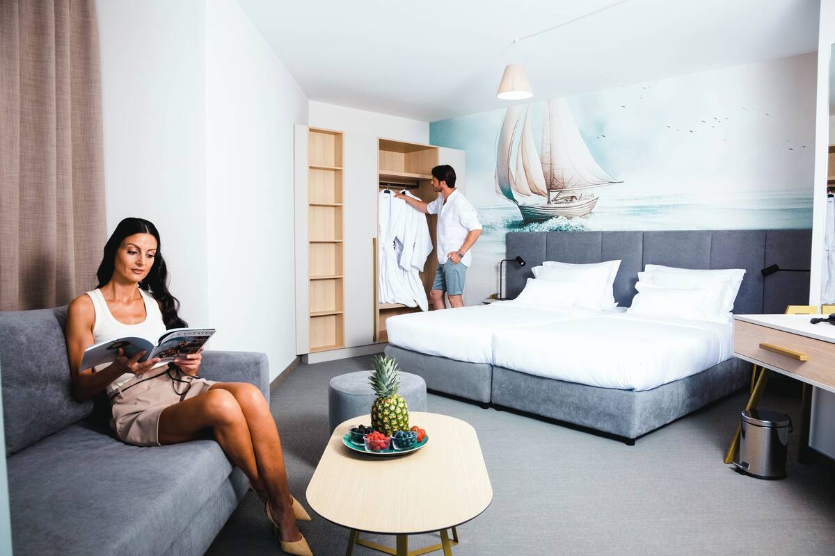 A modern hotel room with a bed, a sofa, a small table with fruit, and a mural of a sailboat.
