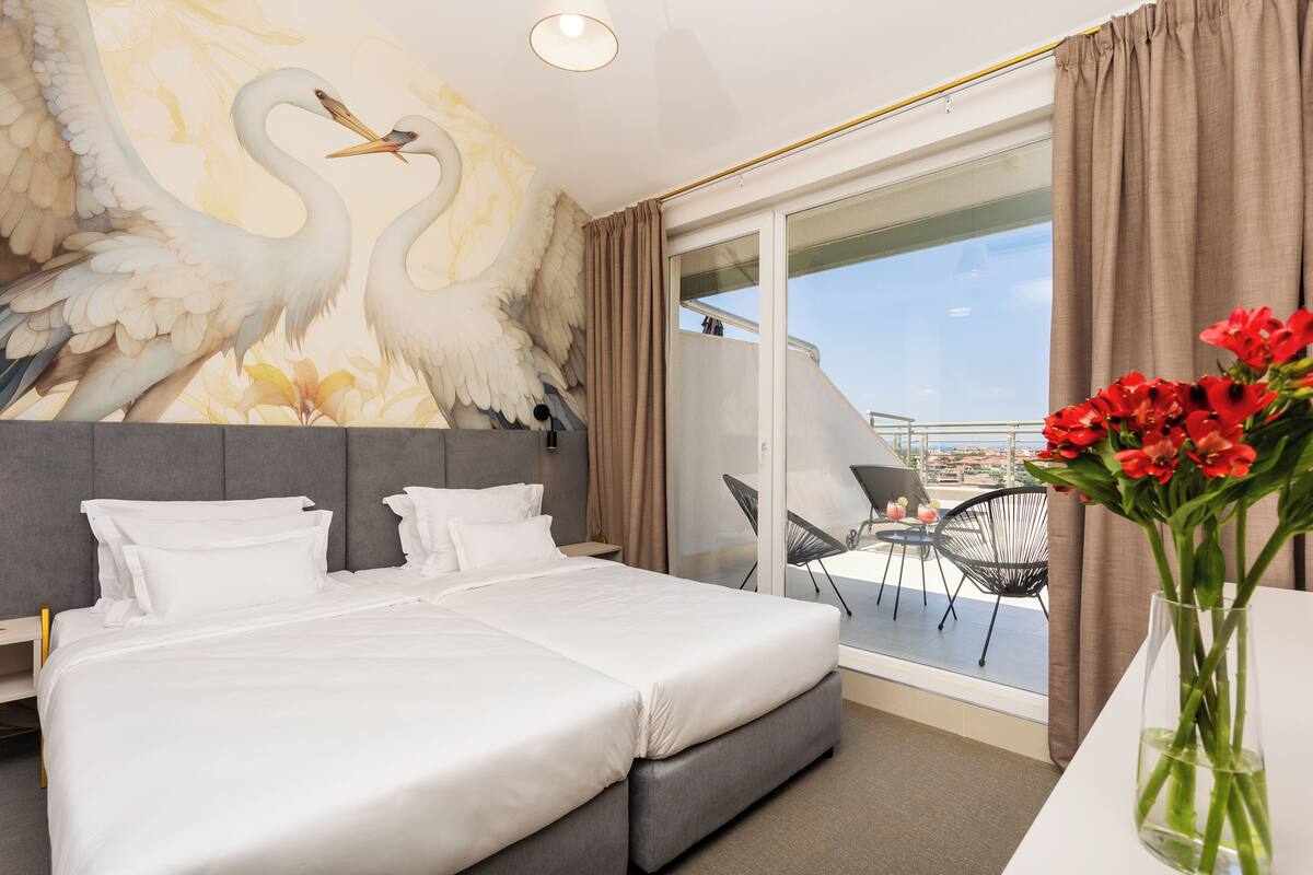 A hotel room with a large bed, a mural of swans, a balcony with a view, and a vase of red flowers.