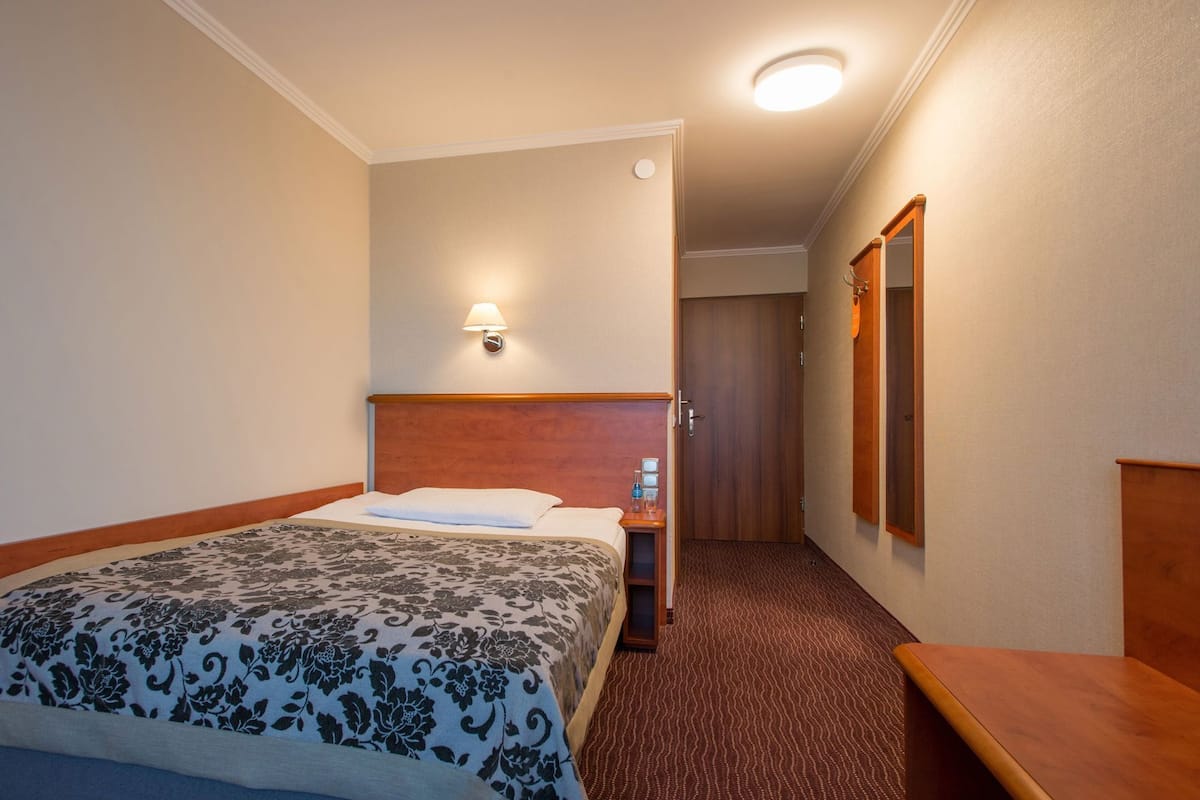A hotel room with a bed, a nightstand with a lamp, a mirror, and a door leading to another room.