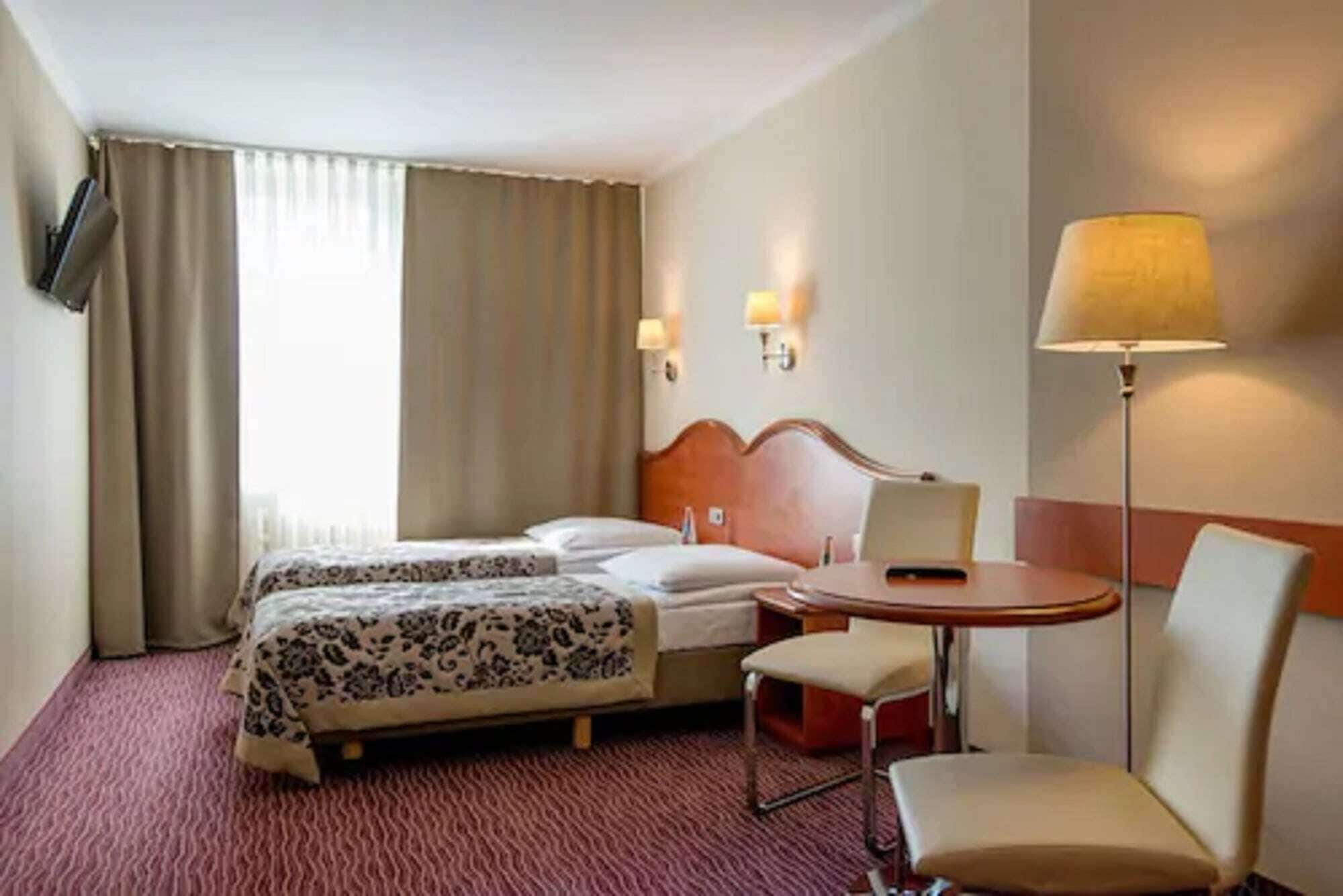 A hotel room with two beds, a desk, a chair, and two wall-mounted lamps.