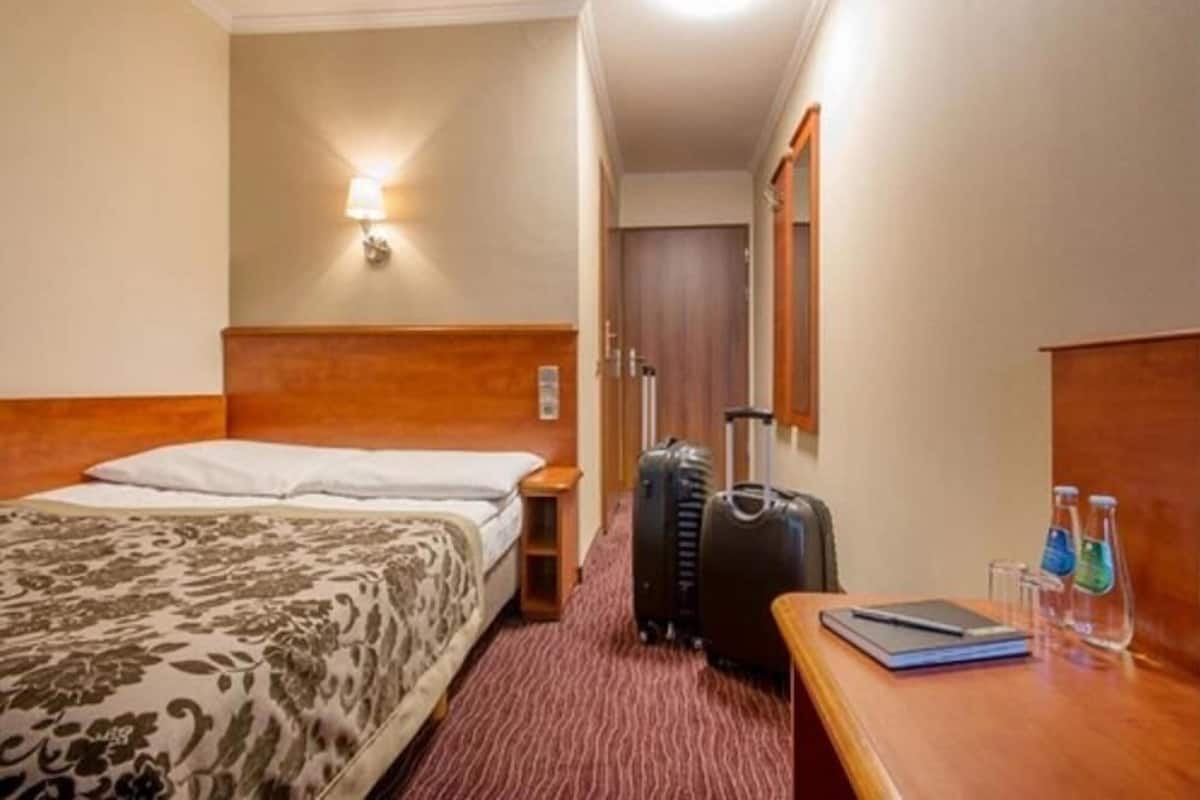 A hotel room with a bed, a desk with a laptop, two suitcases, and a nightstand with a lamp.