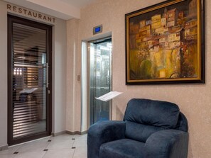 Un hall d’hôtel avec un fauteuil, un tableau, un ascenseur et une enseigne indiquant l’emplacement du restaurant.