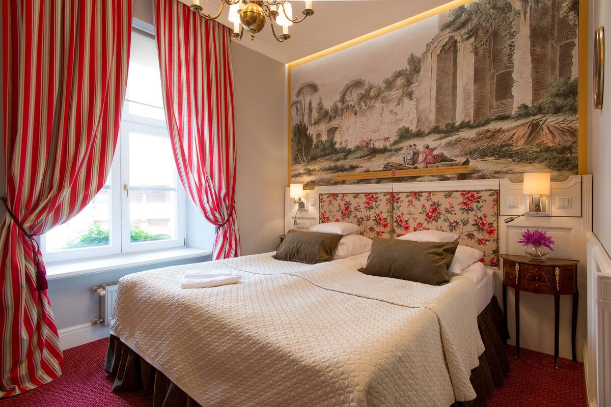 A hotel room with a large bed, red curtains, a floral headboard, and a mural on the wall.