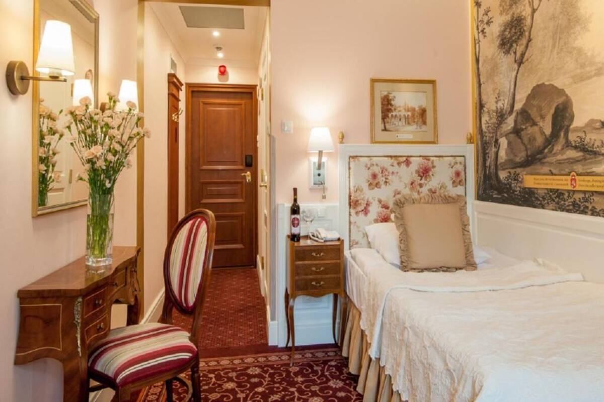 A hotel room with a wooden door, a bed with a floral headboard, a nightstand, a chair, a desk with a lamp, and a vase with flowers.