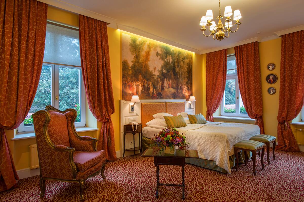 A bedroom with a large bed, a chair, a nightstand, a lamp, and a painting on the wall.