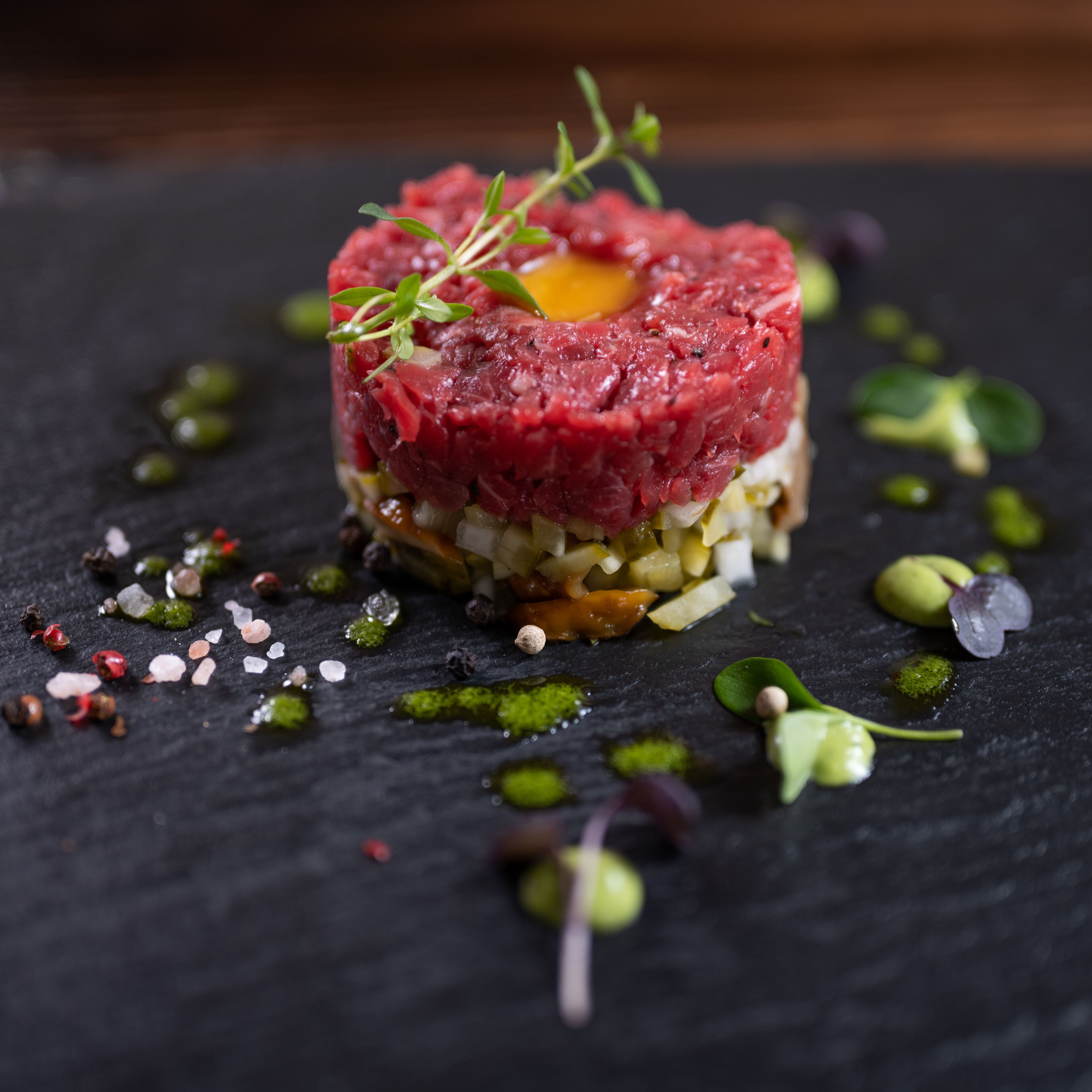 A dish of raw steak tartare with a garnish, surrounded by various condiments and garnishes on a dark slate plate.