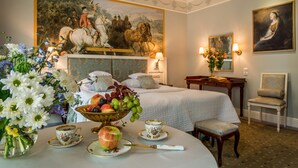 A hotel room with a large bed, a desk with a chair, a painting on the wall, and a dining table with flowers and fruit. - Hotel Polski Pod Białym Orłem (Kraków)