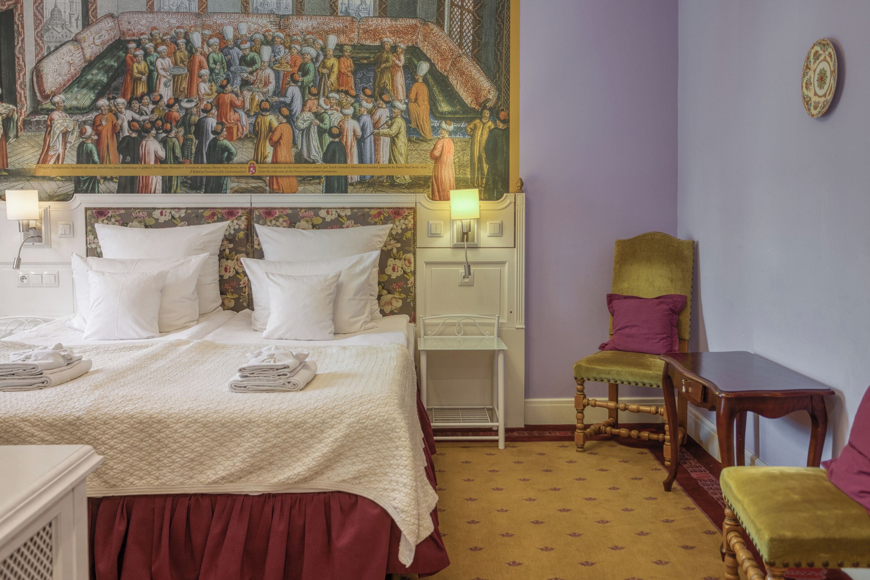 Une chambre d’hôtel avec une grande fresque murale, un lit avec du linge de lit blanc, une table de chevet, une chaise et une petite table.