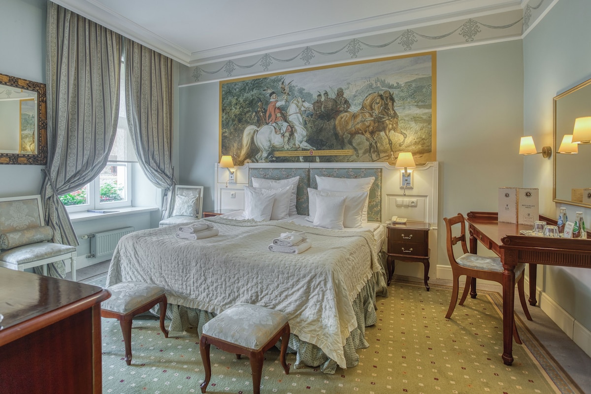 A hotel room with a large bed, a sitting area with a chair and a table, a painting on the wall, and a bathroom with a mirror and a towel rack.