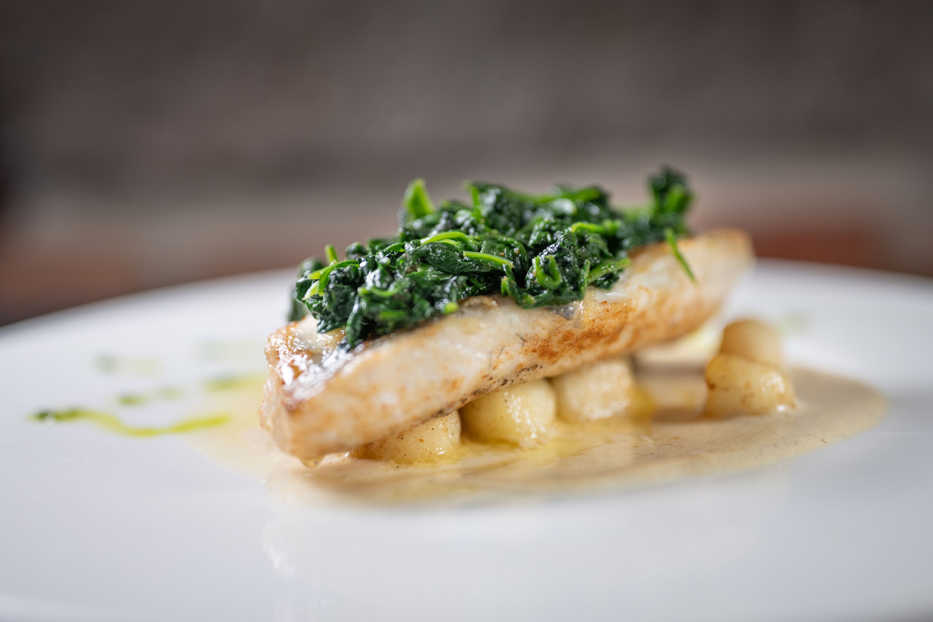 A dish with a piece of fish topped with greens and served with dumplings and sauce.