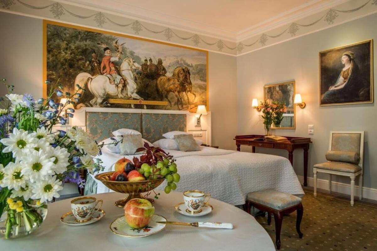 A hotel room with a bed, a desk, a chair, a painting, and a fruit bowl.