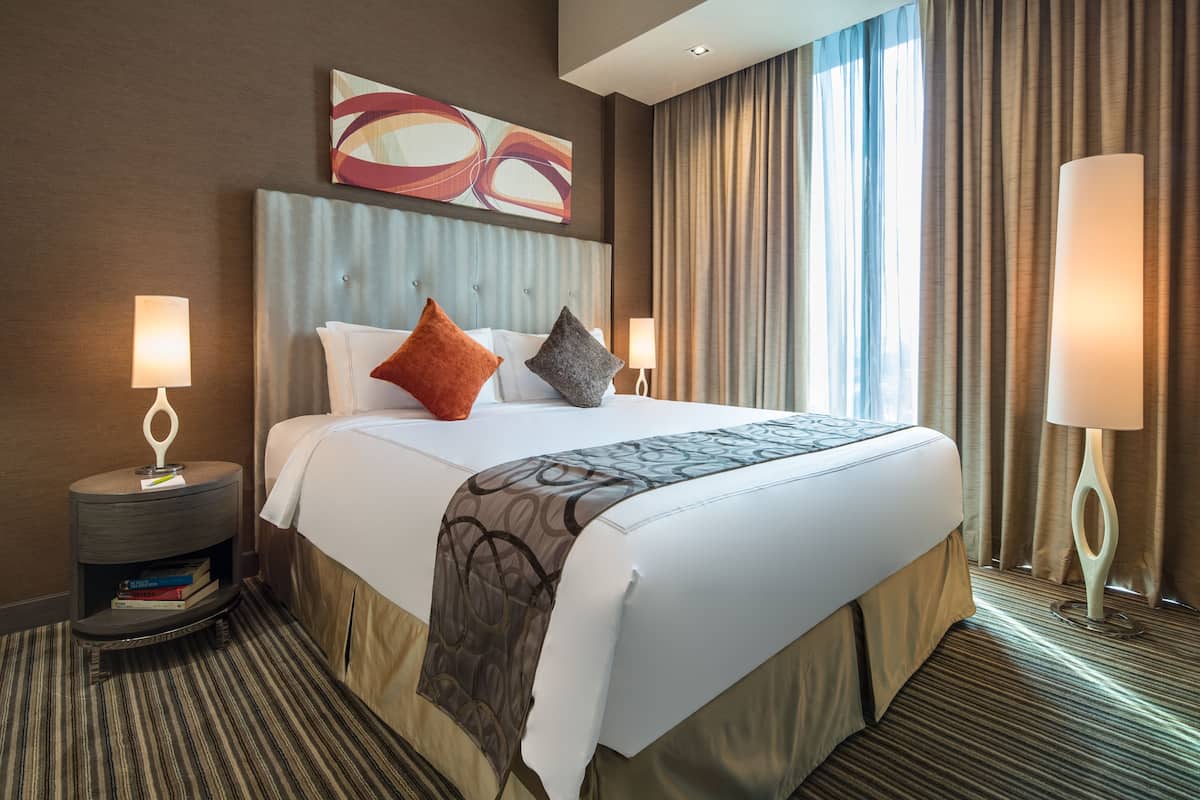 A hotel room with a large bed, two bedside tables with lamps, a headboard, and a window with curtains.