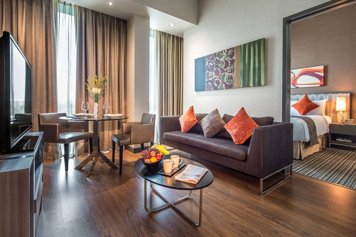 A modern hotel room with a sofa, a dining area, a TV, and a large window with curtains.
