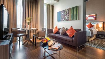 A modern hotel room with a sofa, a dining area, a TV, and a large window with curtains.