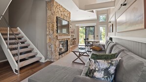 Una sala de estar moderna con una pared de acento de piedra, una escalera, un televisor de pantalla plana y un comedor con luz natural.