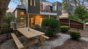 Una casa moderna de dos pisos con un patio, una mesa de madera y un pequeño jardín.
