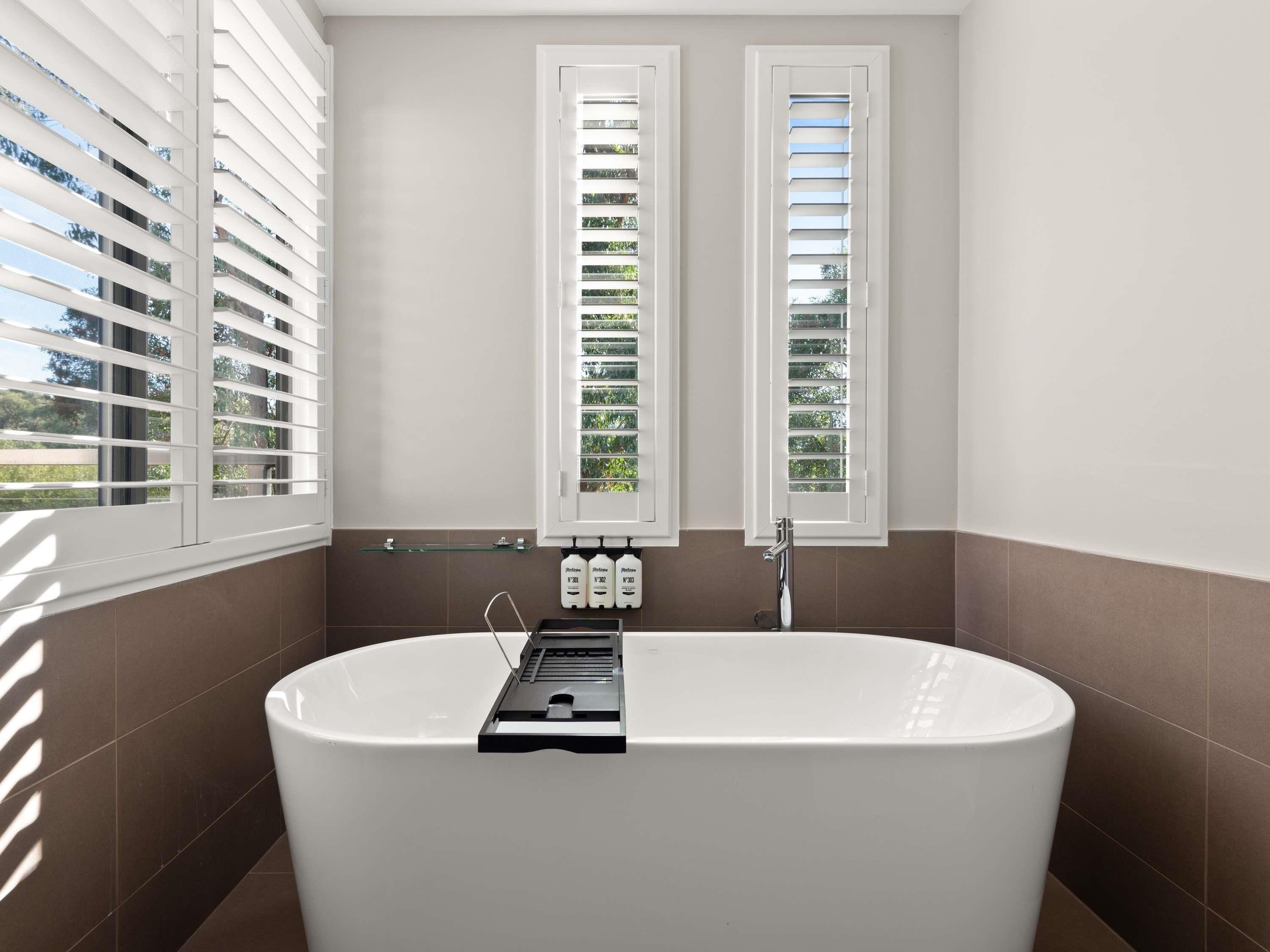 A modern bathroom with a freestanding bathtub, two windows with blinds, and a glass shelf with toiletries.