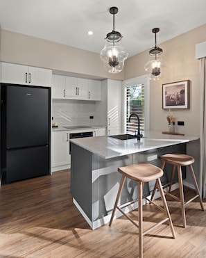Una cocina moderna con armarios blancos, un refrigerador negro, una isla central con taburetes, iluminación colgante y un cuadro enmarcado en la pared.
