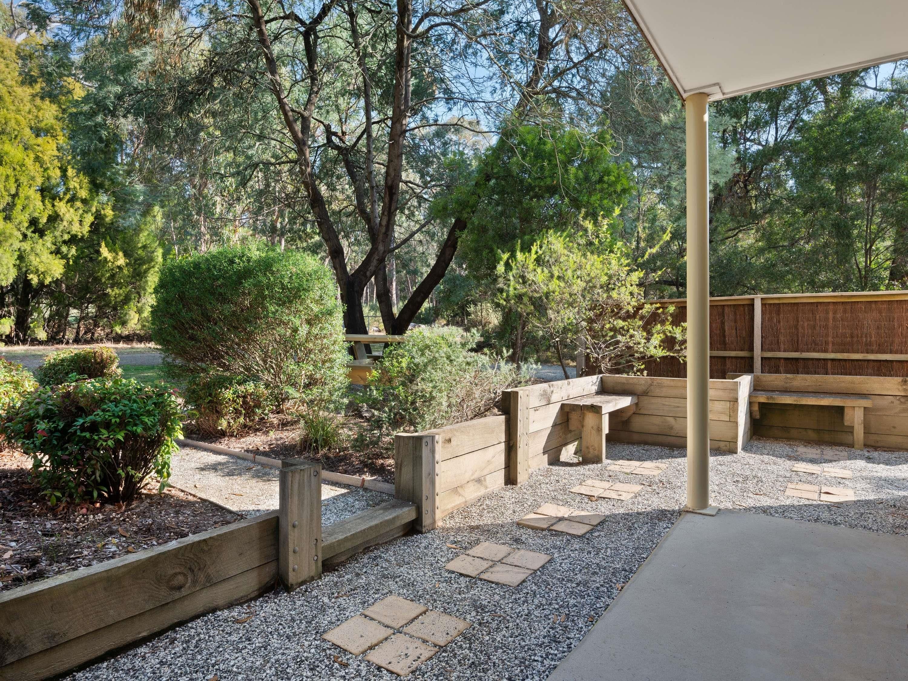 A covered outdoor seating area with wooden benches and a pergola, surrounded by gravel and small plants.