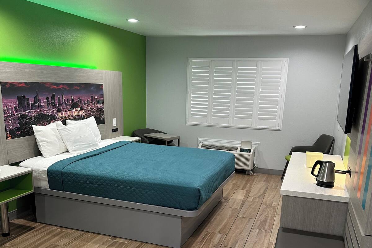 Quarto de hotel moderno com cama, mesas de cabeceira, escrivaninha, cadeira, televisão e janela com persianas.