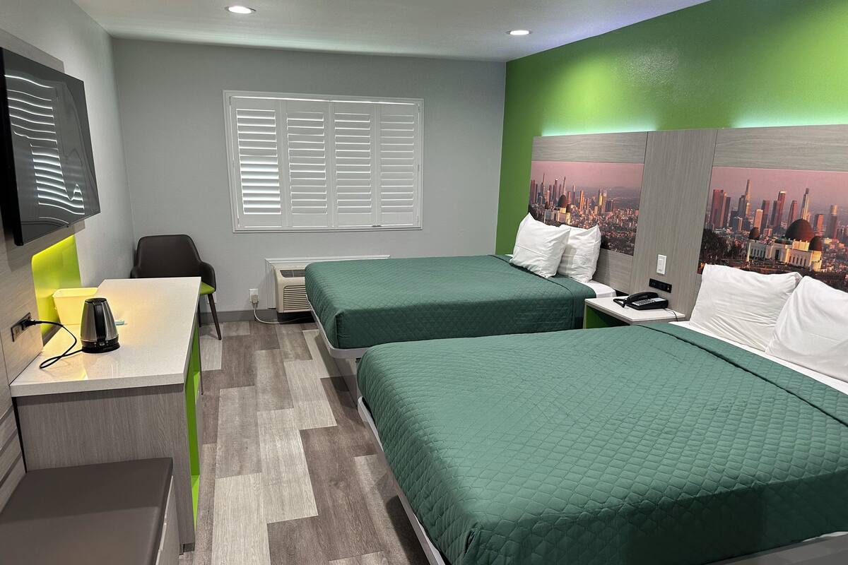 Quarto de hotel com duas camas, uma escrivaninha com luminária, uma cadeira, uma janela com persianas e um mural com vista para a cidade na parede.