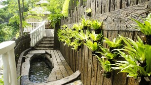 Eine Holztreppe führt zu einem Gebäude mit einem Strohdach, umgeben von üppiger Vegetation und einem kleinen Wasserspiel.