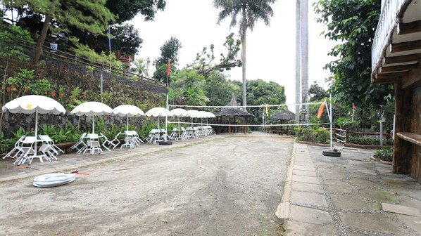 An outdoor seating area with white tables and chairs under white umbrellas, surrounded by greenery and a stone wall.