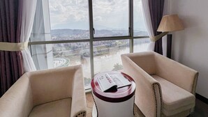 A hotel room with a large window, two beige armchairs, a small round table with a magazine, and a lamp.