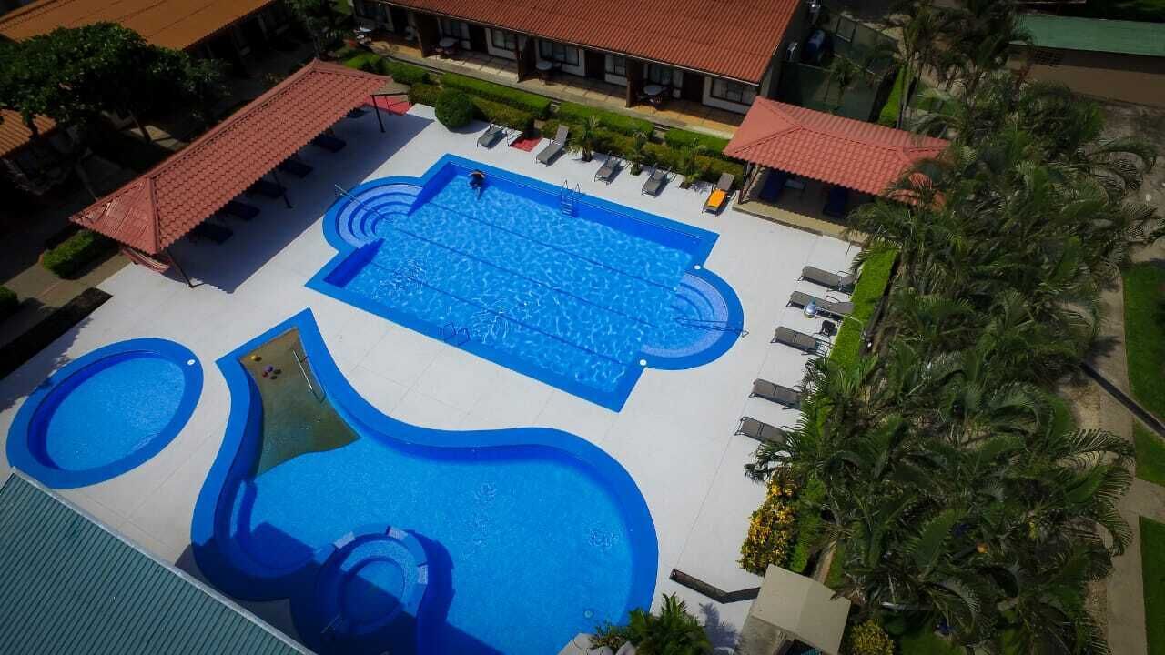 Vista aérea de un hotel con una piscina central grande, piscinas más pequeñas alrededor y un área de estar cubierta con sombrillas.