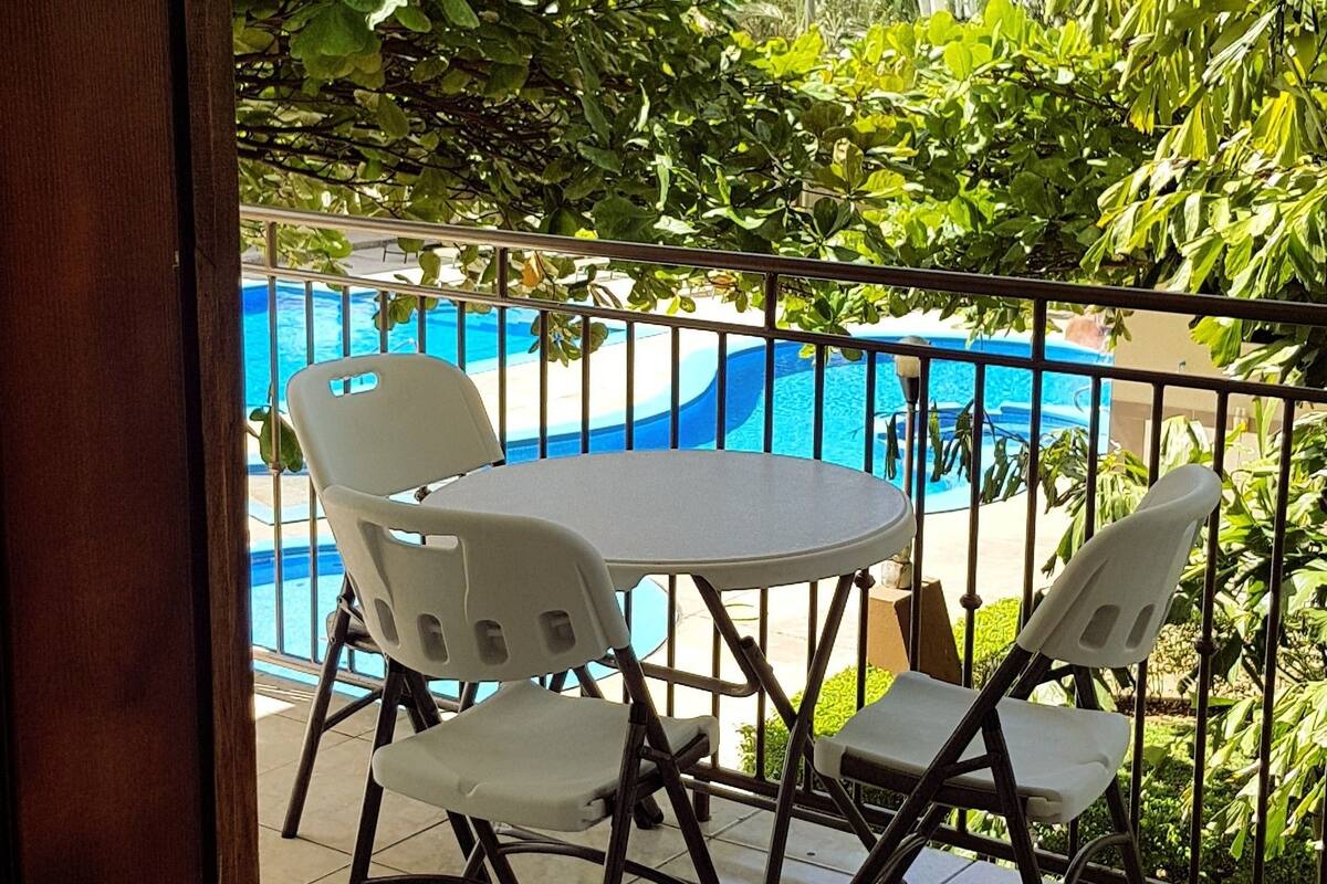 A balcony with a table and chairs, overlooking a swimming pool and lush greenery.