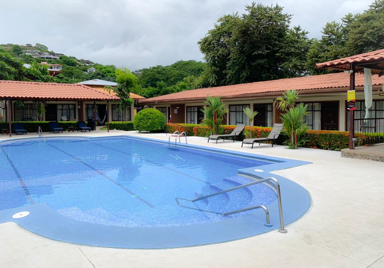 A swimming pool with lounge chairs and a covered seating area, surrounded by greenery and buildings with red-tiled roofs.