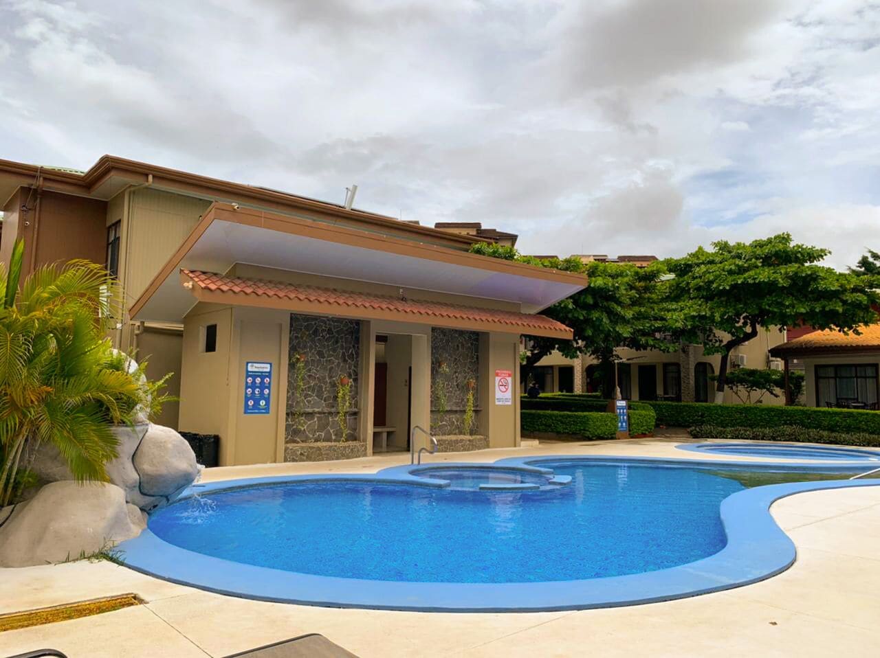 A swimming pool with a stone wall and a covered area, surrounded by greenery and a building with a tiled roof.