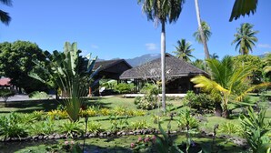 A tropical garden with a pond, palm trees, and a building with a thatched roof. - Hotel Royal Tahitien (Pirae)