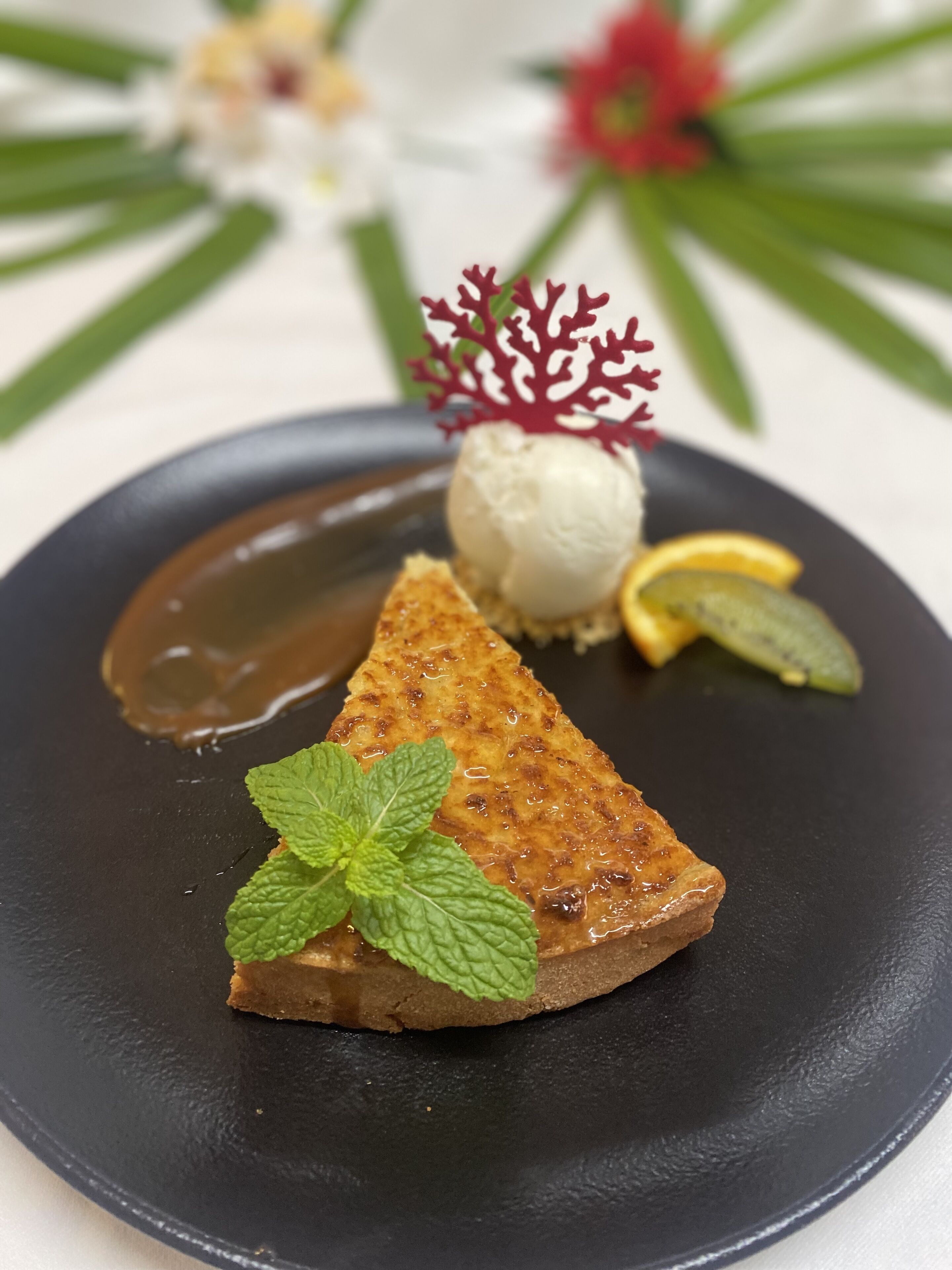 A dessert plate with a slice of cake, a scoop of ice cream, a mint leaf, and a decorative red coral piece.
