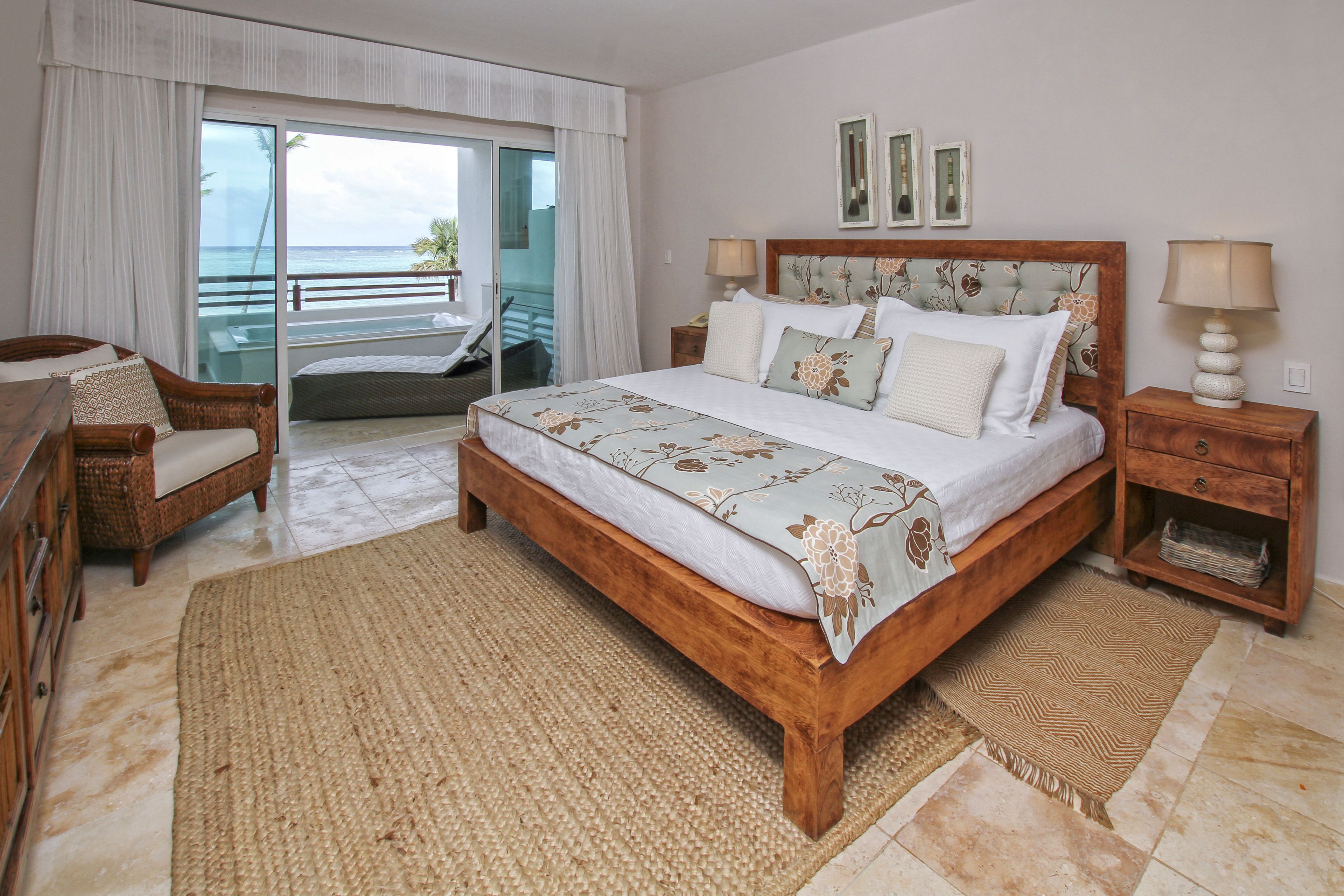 A bedroom with a large bed, a wooden nightstand, a wicker chair, a balcony with a view of the ocean, and a sliding glass door.