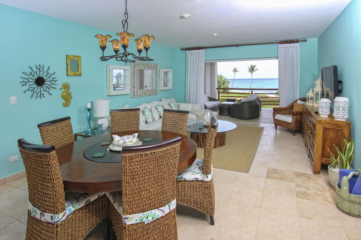 A dining area with wicker chairs and a wooden table, a living room with a sofa and a TV, and a view of the ocean through large windows.
