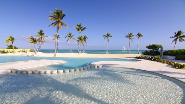 Una piscina azul cristalina rodeada de arena blanca y palmeras, con vista al océano y un velero a lo lejos.