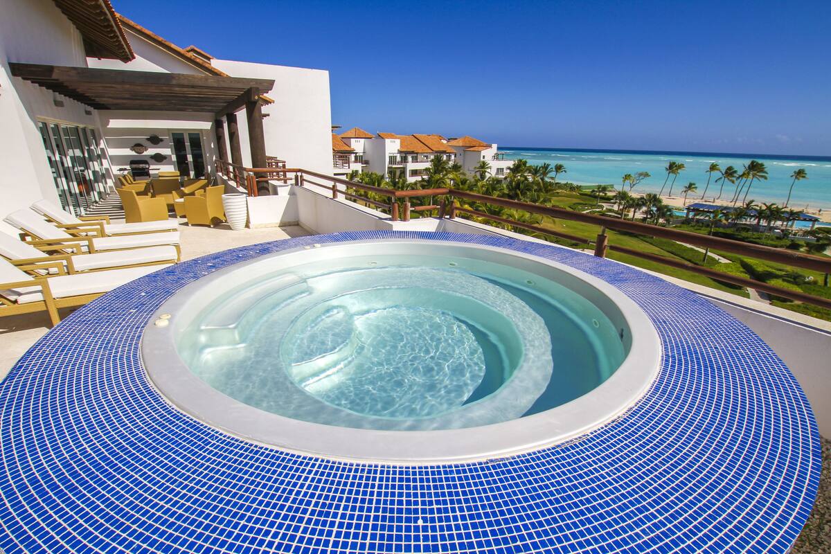 A hot tub with a tiled edge, a poolside area with lounge chairs, and a view of the ocean and resort buildings.