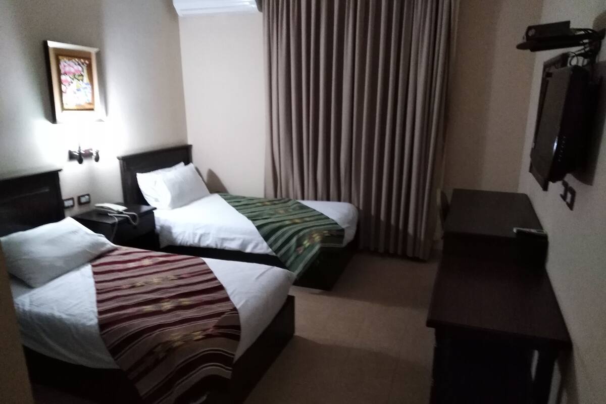 A hotel room with two beds, a desk, a chair, a TV, and a window with curtains.