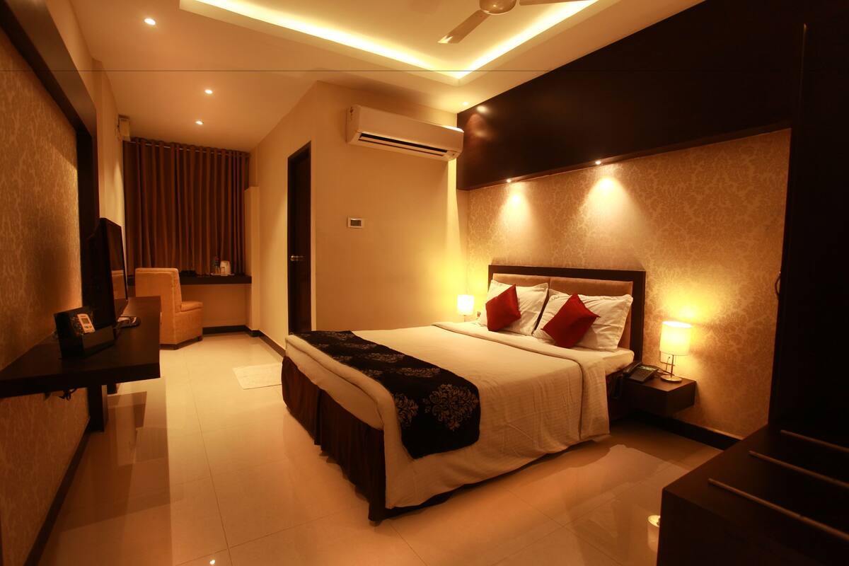 A hotel room with a bed, bedside table, lamp, and an air conditioning unit.