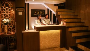 A hotel reception desk with two staff members, a staircase, and a decorative wall with a sign for free airport transfers. - Hotel Mars Classic (Chennai)