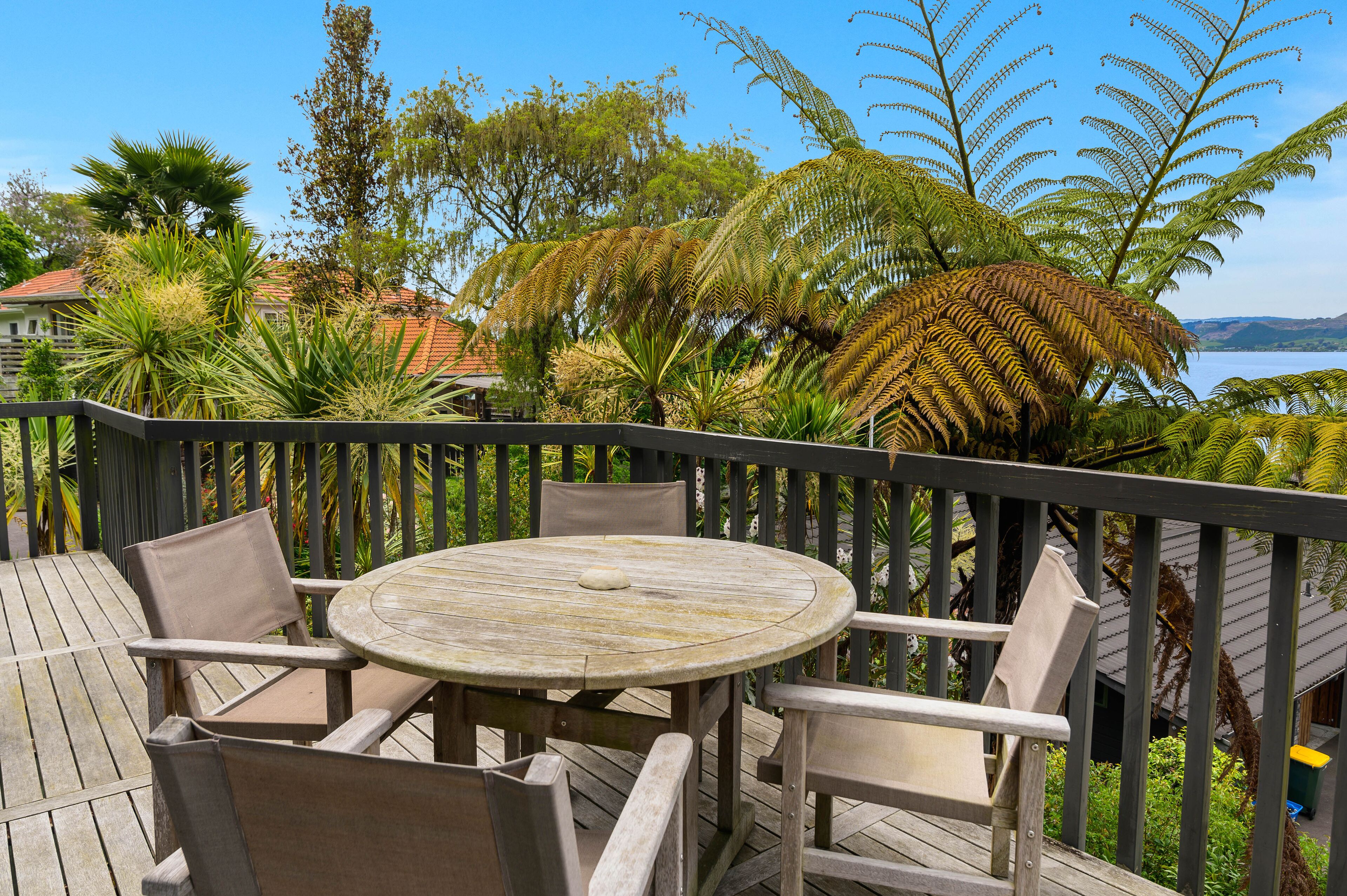 Una terraza de madera con una mesa redonda y sillas, rodeada de abundante follaje y vistas a un cuerpo de agua.