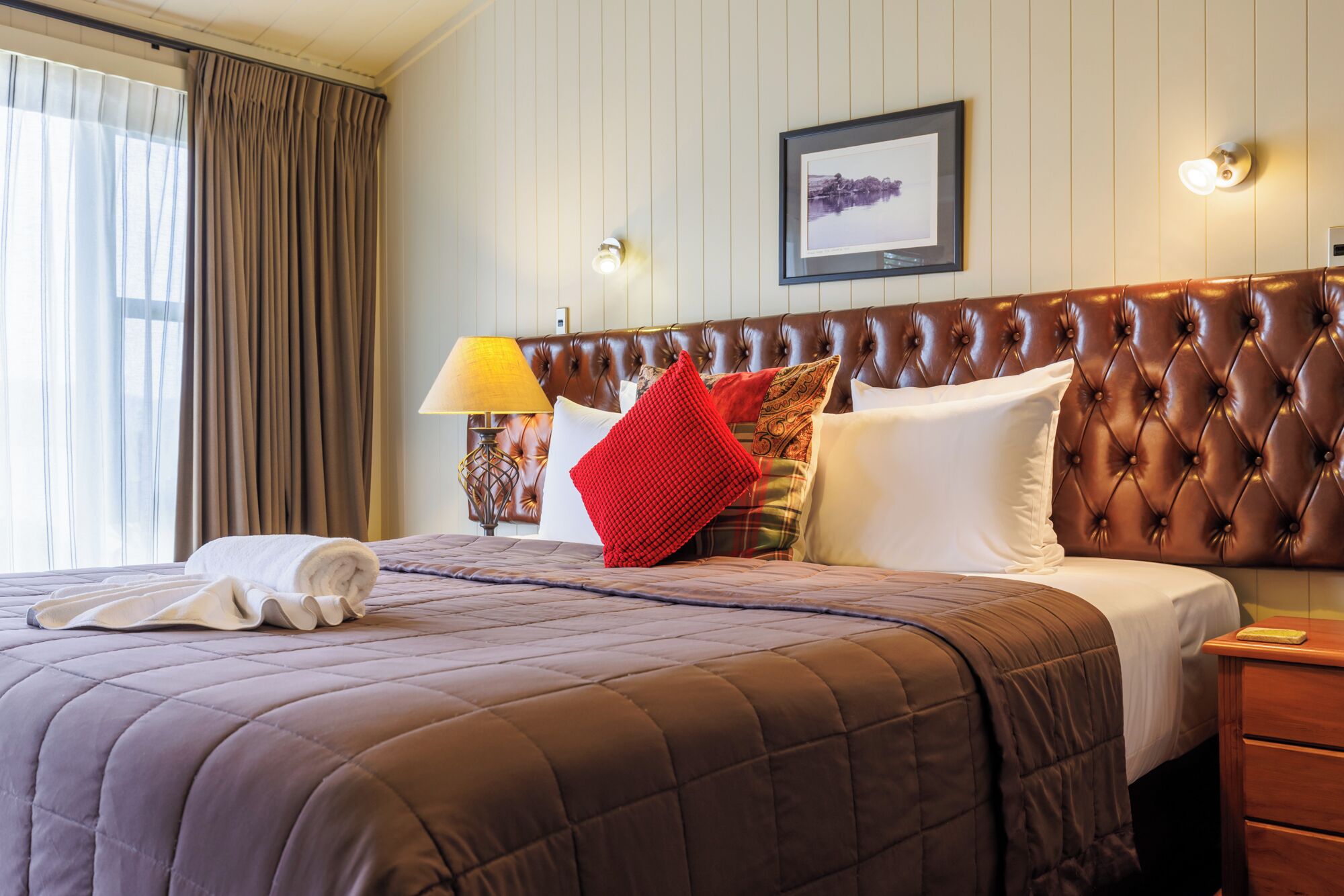 A hotel room with a large bed, a headboard, a bedside table, a lamp, and a framed picture on the wall.
