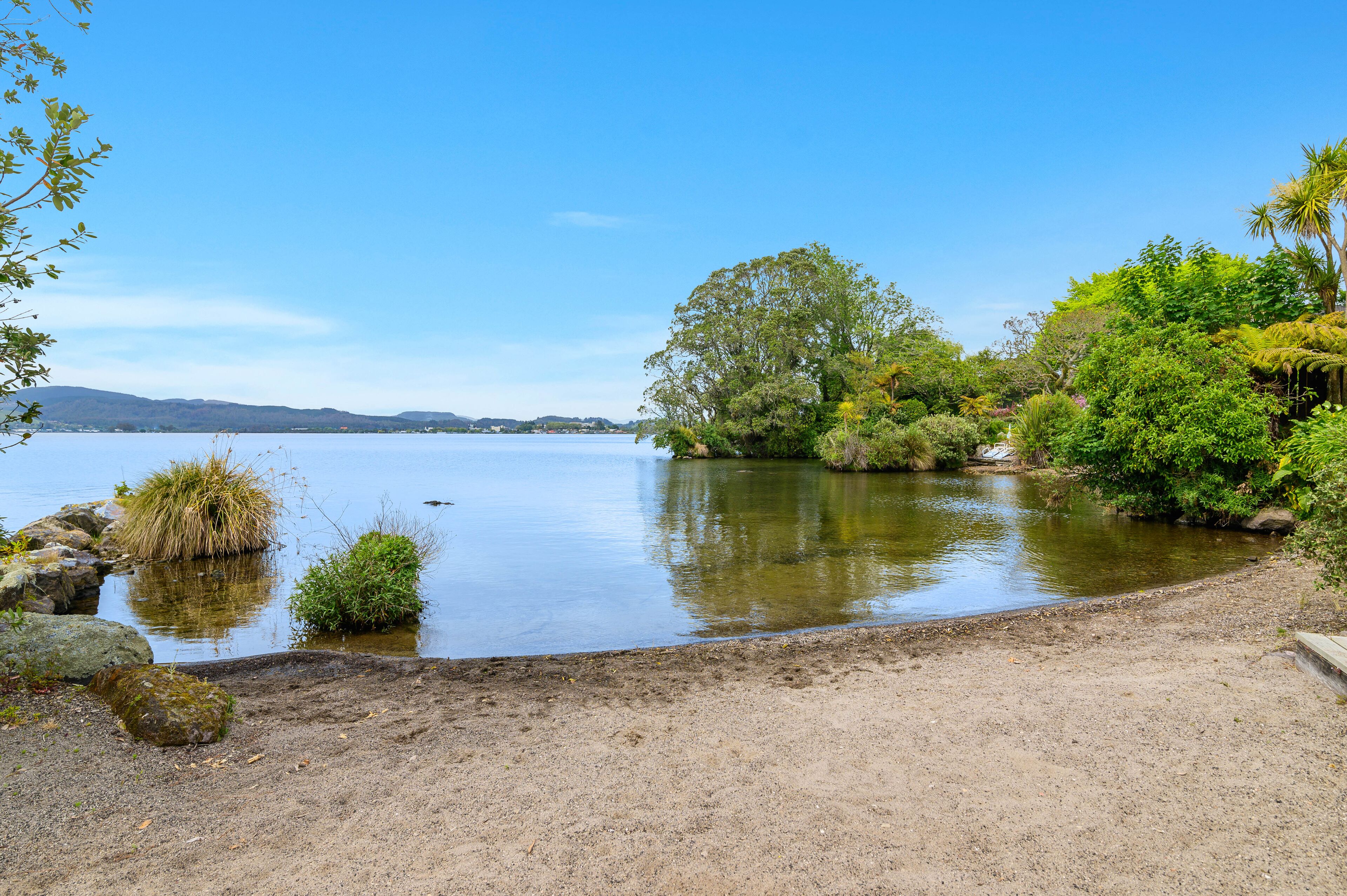 A serene lakeside setting with a sandy shore, dense greenery, and calm water reflecting the sky.
