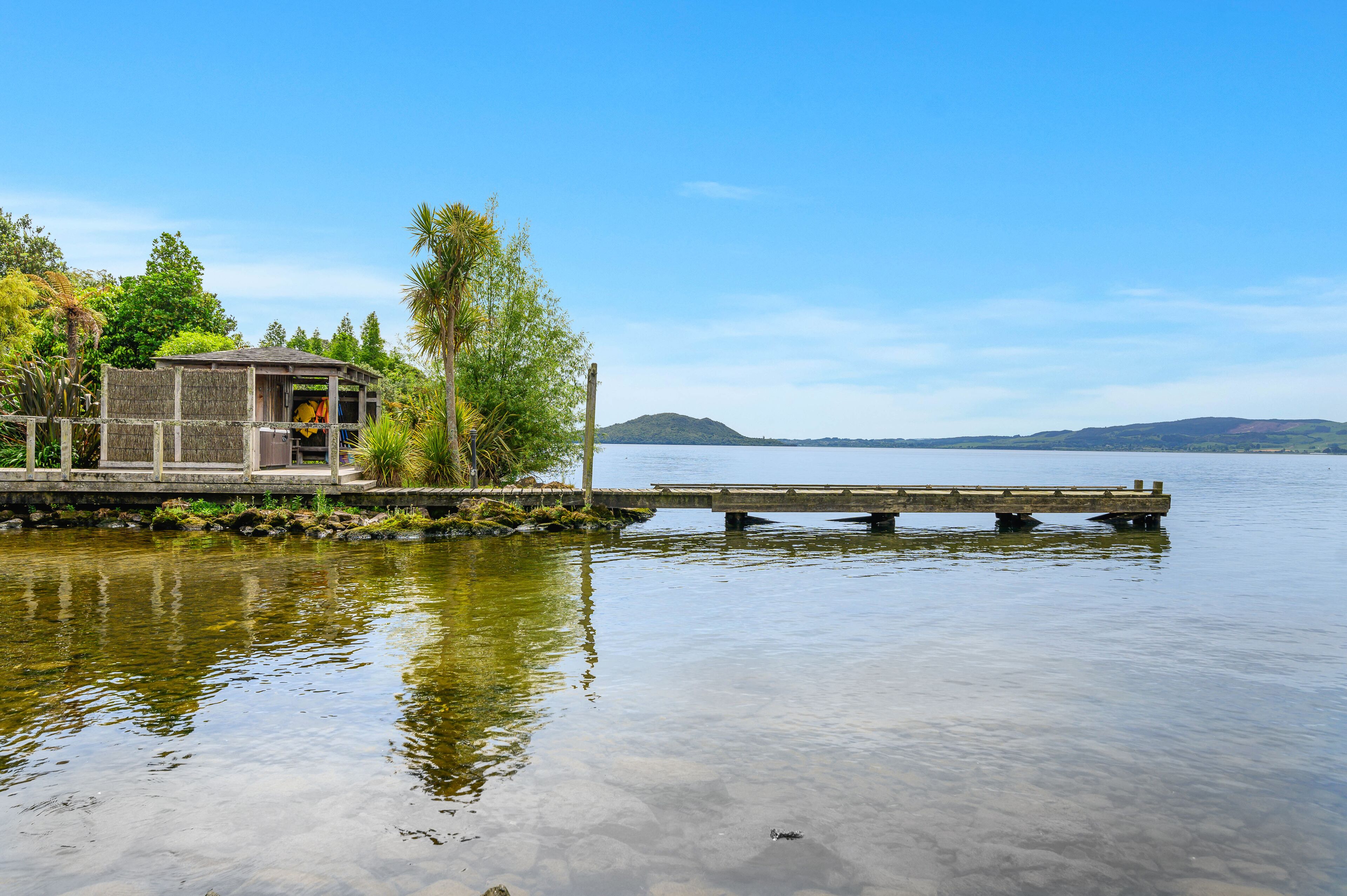 Un lago tranquilo con un muelle de madera que se adentra en el agua. Se ve una pequeña estructura de madera con techo en el lado izquierdo del muelle.