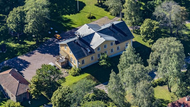 Luchtbeeld van een groot, geel gebouw met zonnepanelen op het dak, omringd door weelderige begroeiing en bomen.