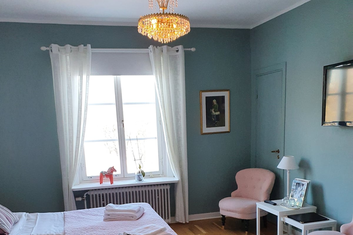 Een slaapkamer met een bed, een stoel, een tafel, een lamp en een raam met gordijnen.