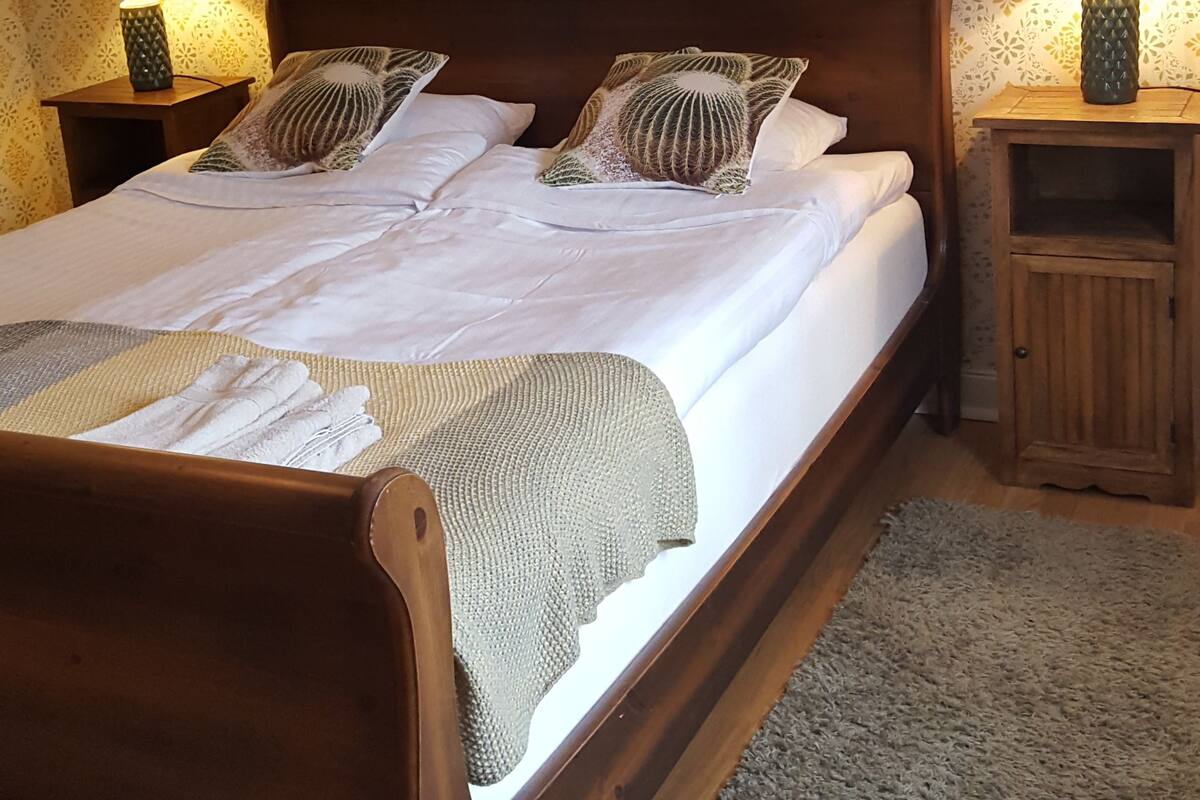 Een tweepersoonsbed met houten frame, twee nachtkastjes met lampen, behang met patroon en een grijze vloerkleed.